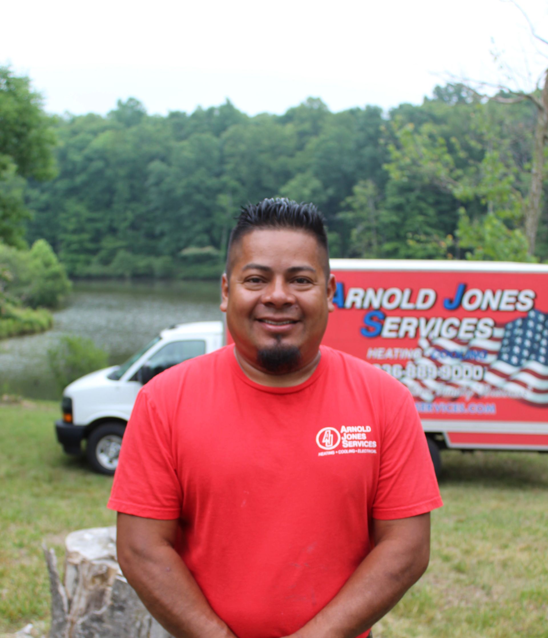Man in red T-shirt smiling outdoors, with a service truck and wooded lake background.