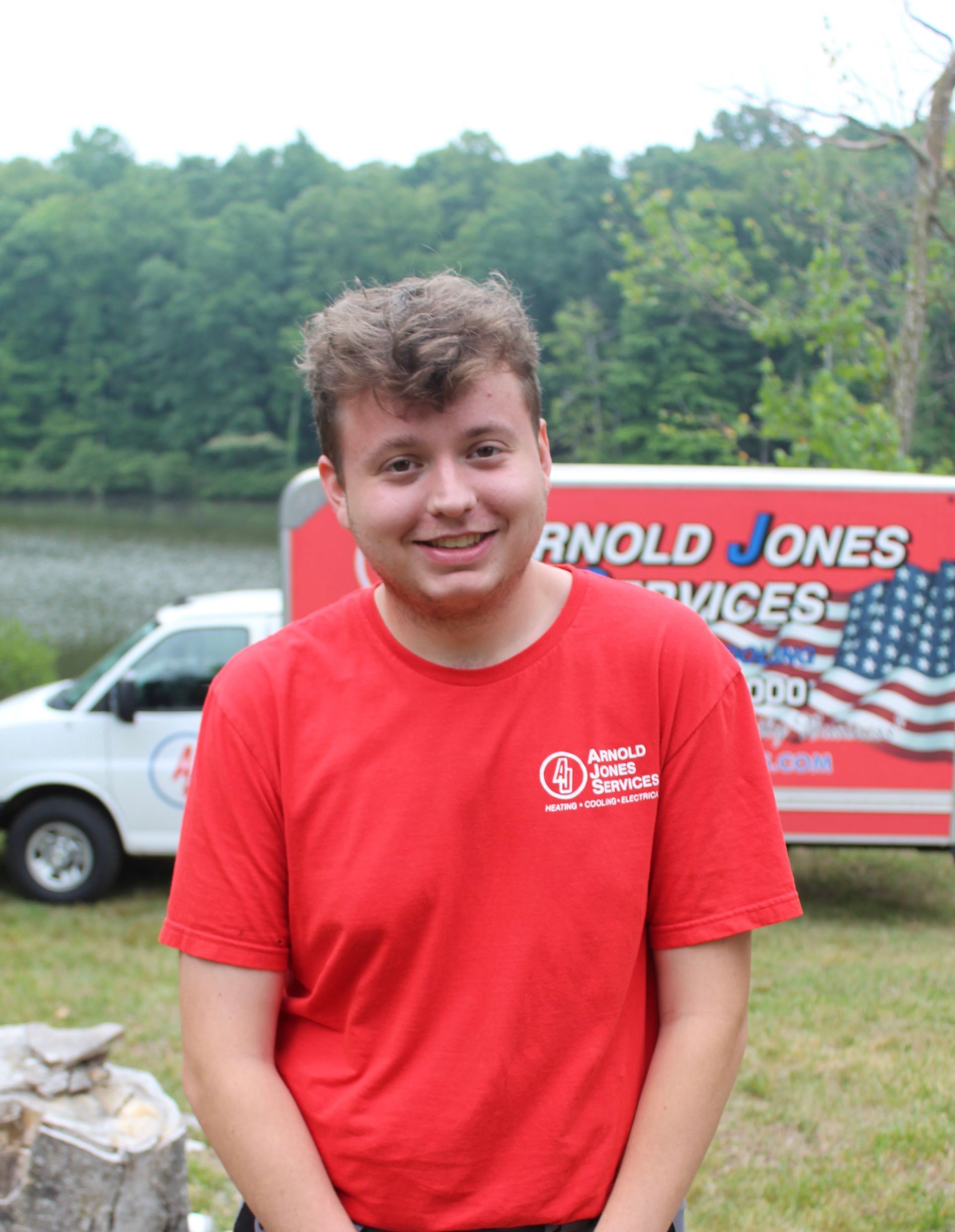 Smiling person in a red T-shirt standing outdoors in front of a truck and trees