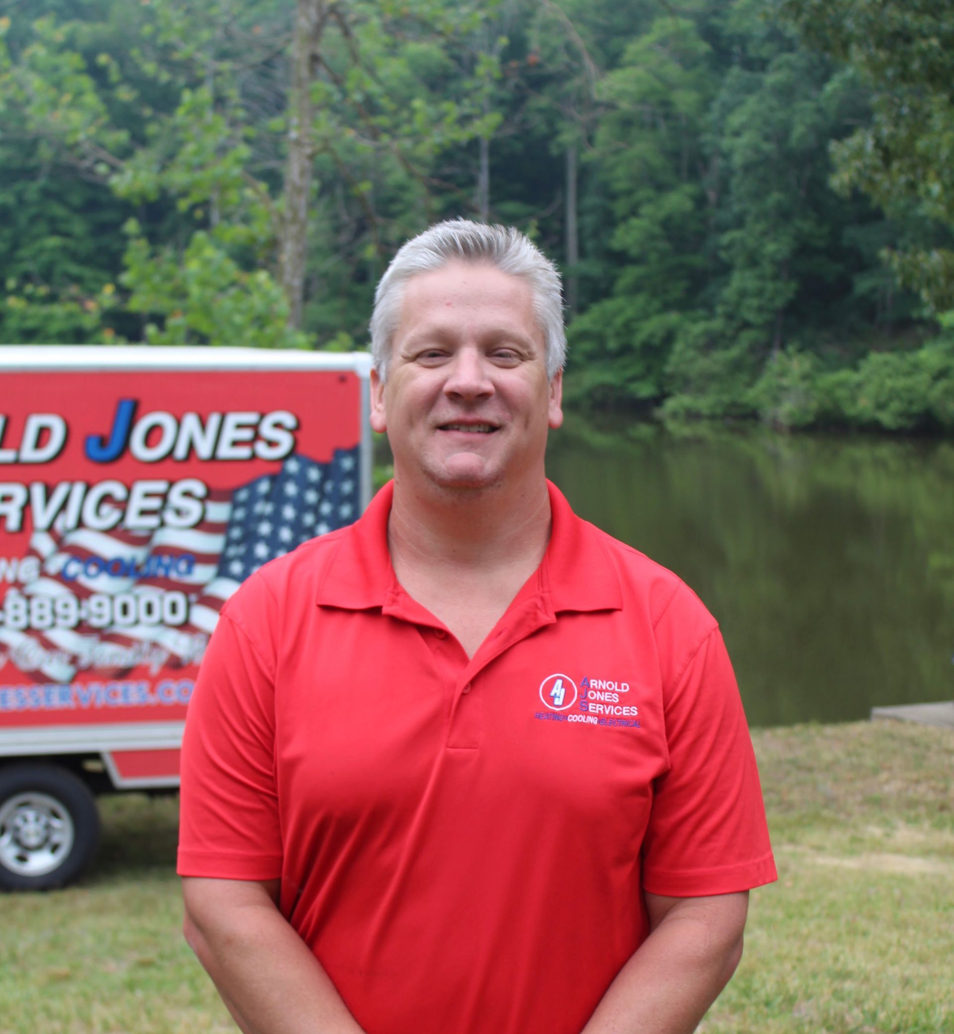 Man in a red polo stands outdoors by a pond, with a service trailer behind him.