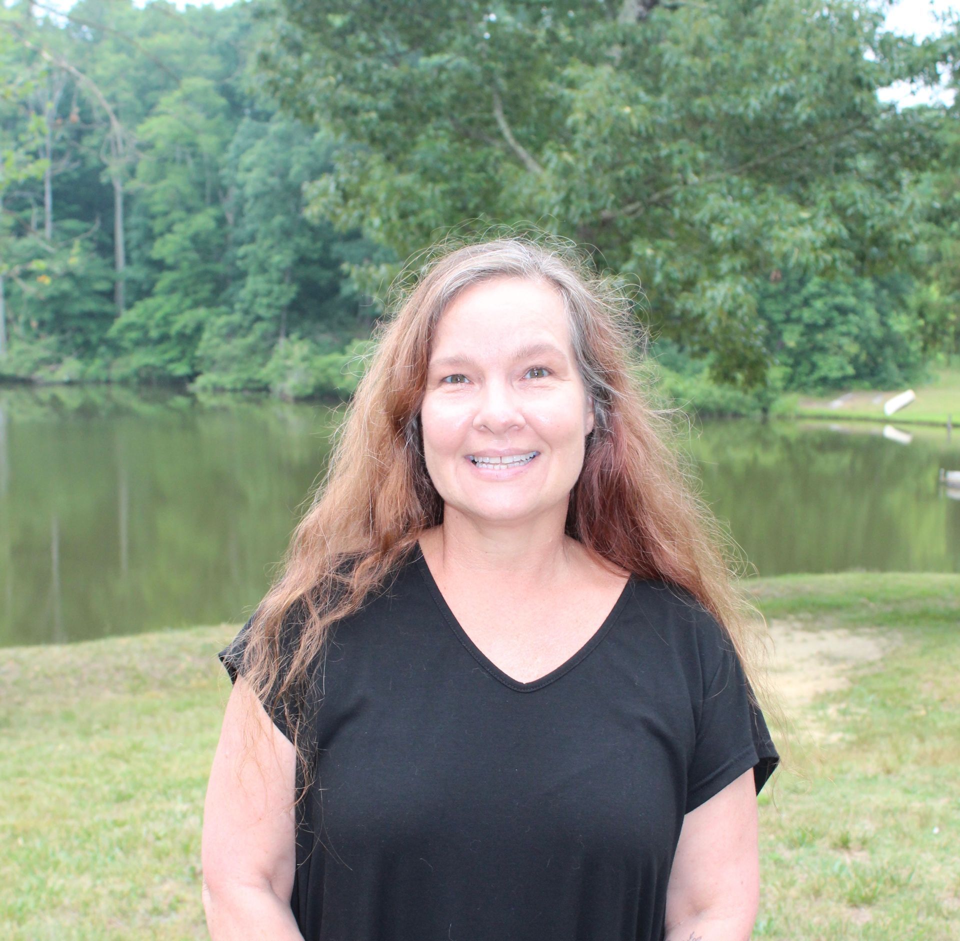 Smiling woman in a black shirt standing by a pond with trees in the background