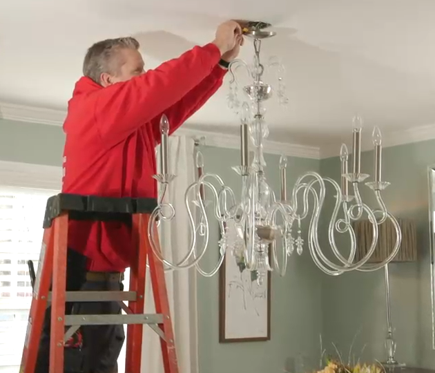 Person in a red shirt installing a chandelier on a ceiling ladder indoors