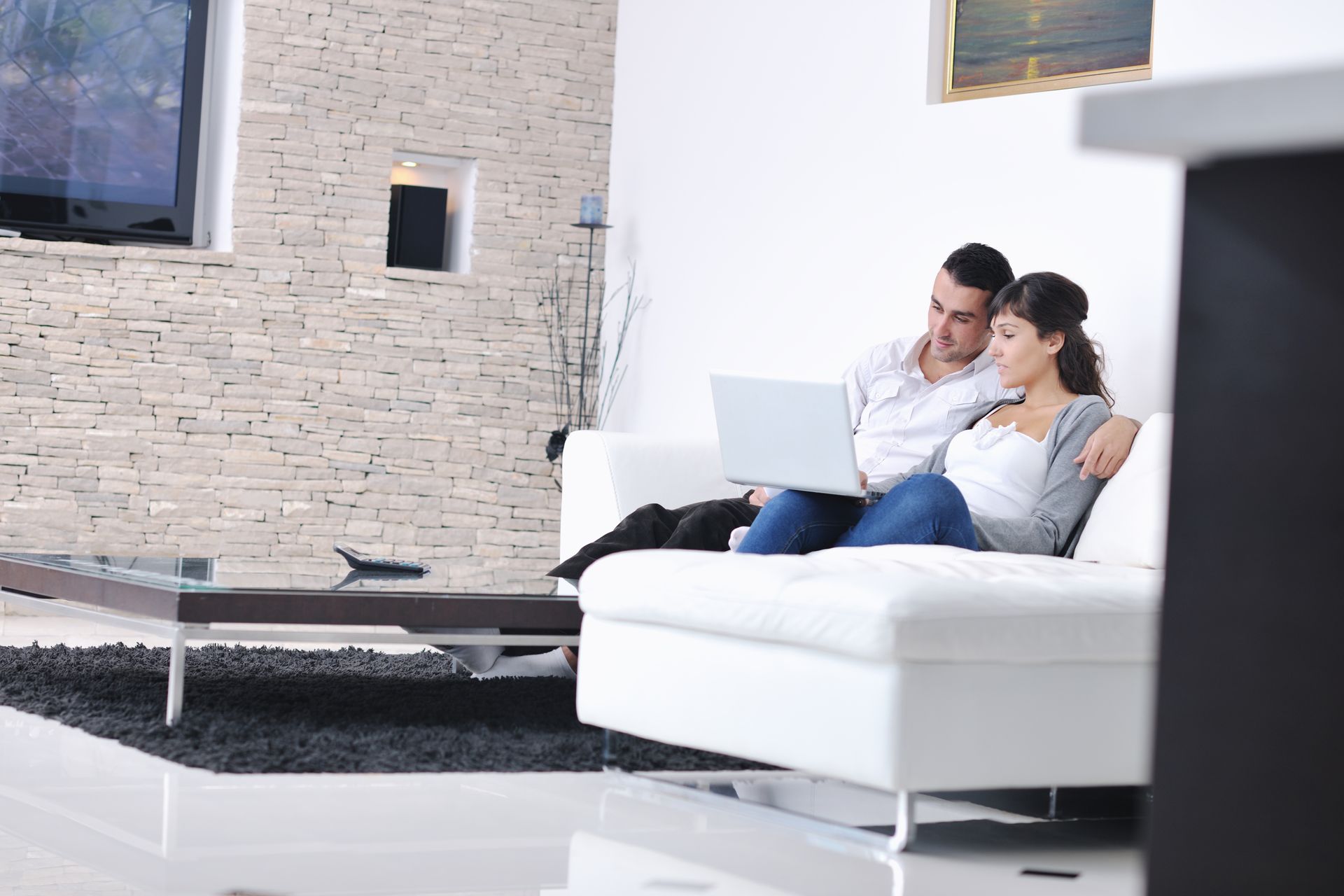 Couple sitting on a white sofa using a laptop in a modern living room with a TV and coffee table.