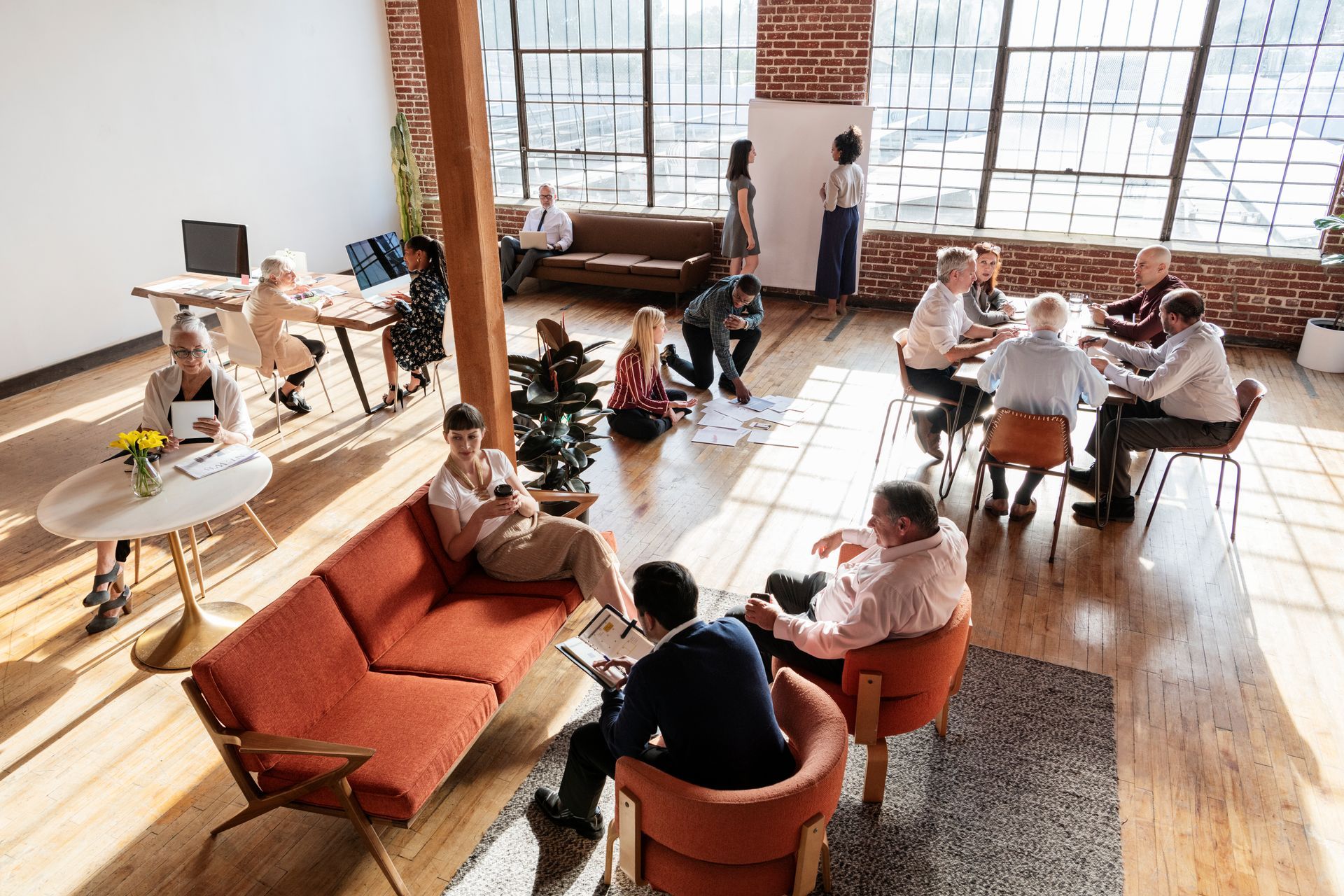 People seated in a bright lounge with large windows, couches, and round tables, socializing and working