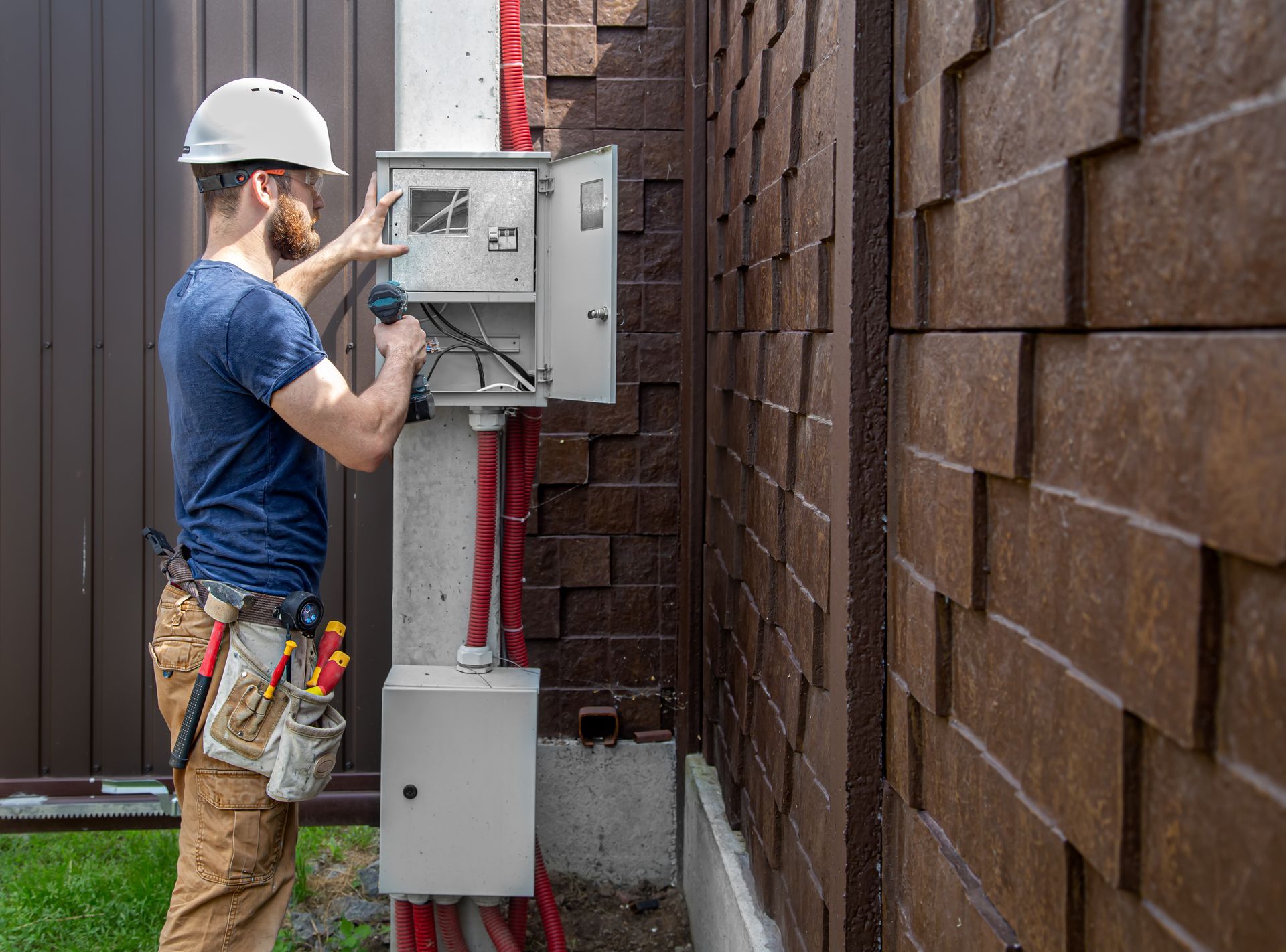 Technician in hard hat working on an outdoor electrical panel beside a brown wall