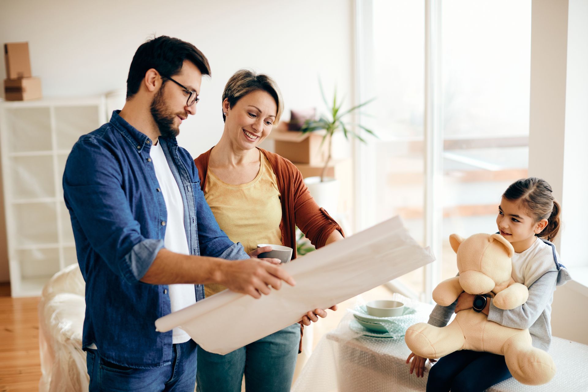 Three people review blueprints in a bright living room; one child holds a teddy bear.