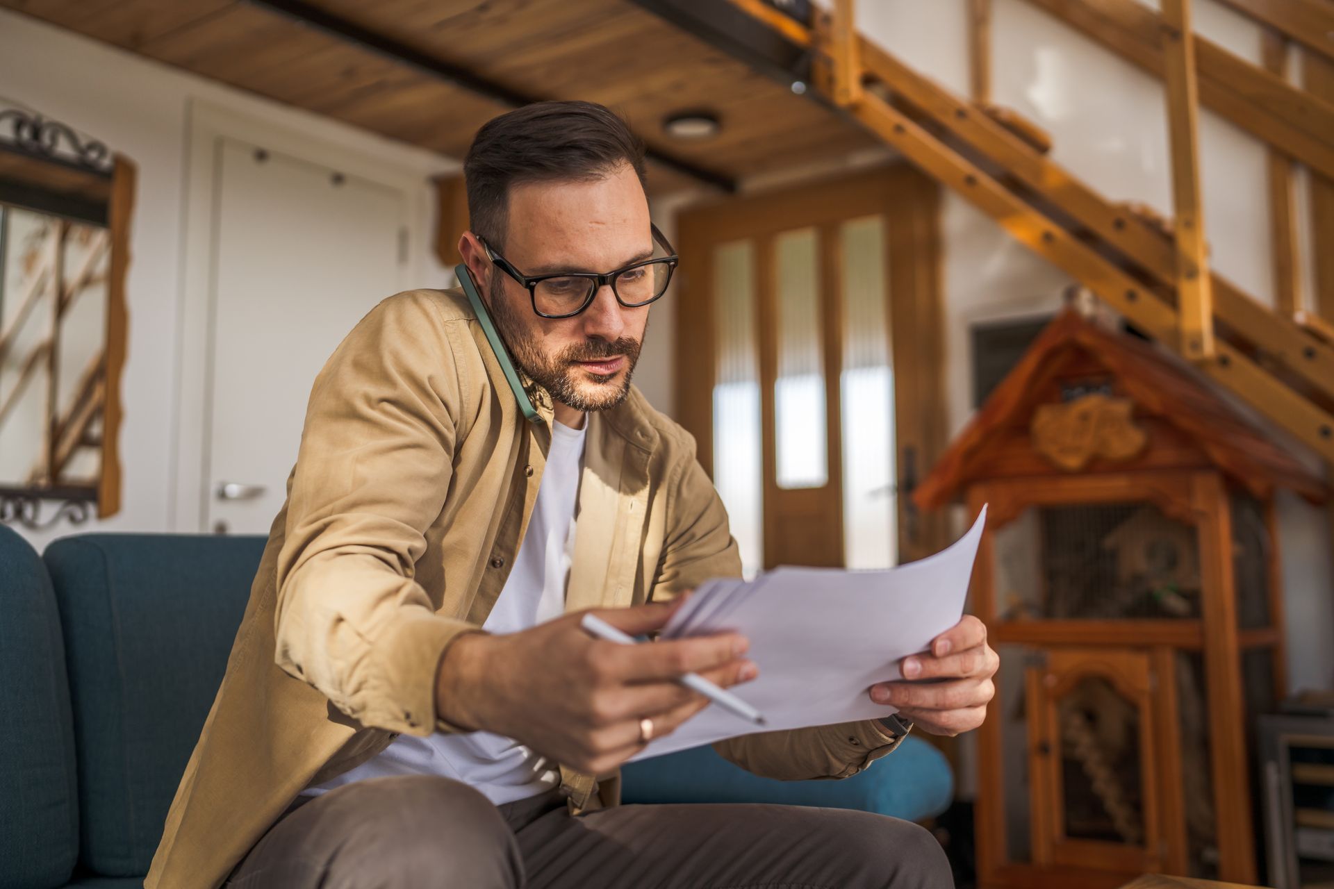 Man reading papers on a couch in a cozy loft with wooden stairs and a small house-shaped shelf
