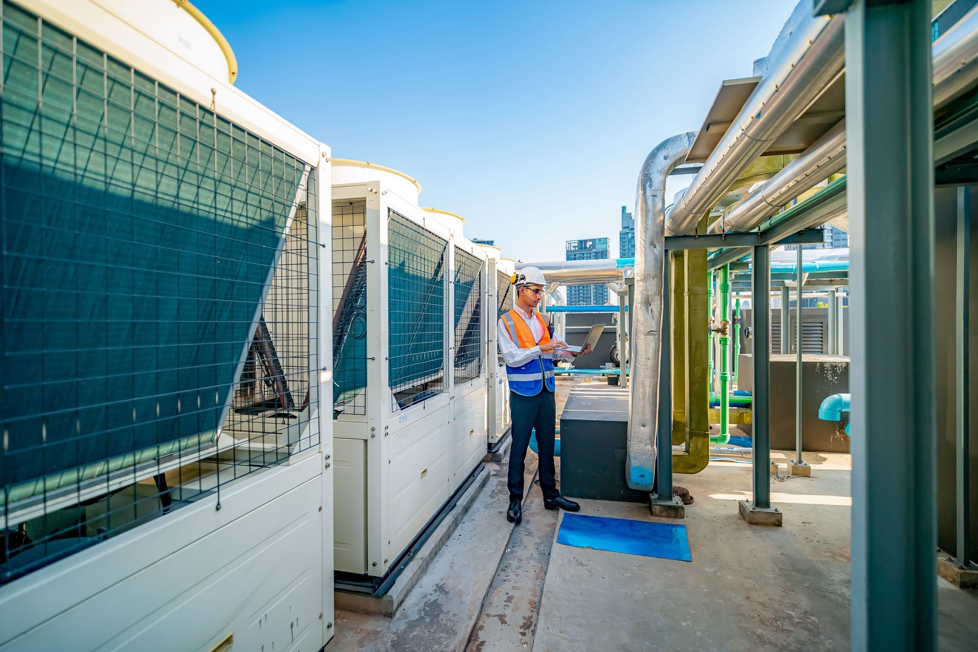 Worker standing between industrial HVAC units and pipes on a rooftop walkway, wearing a safety vest.