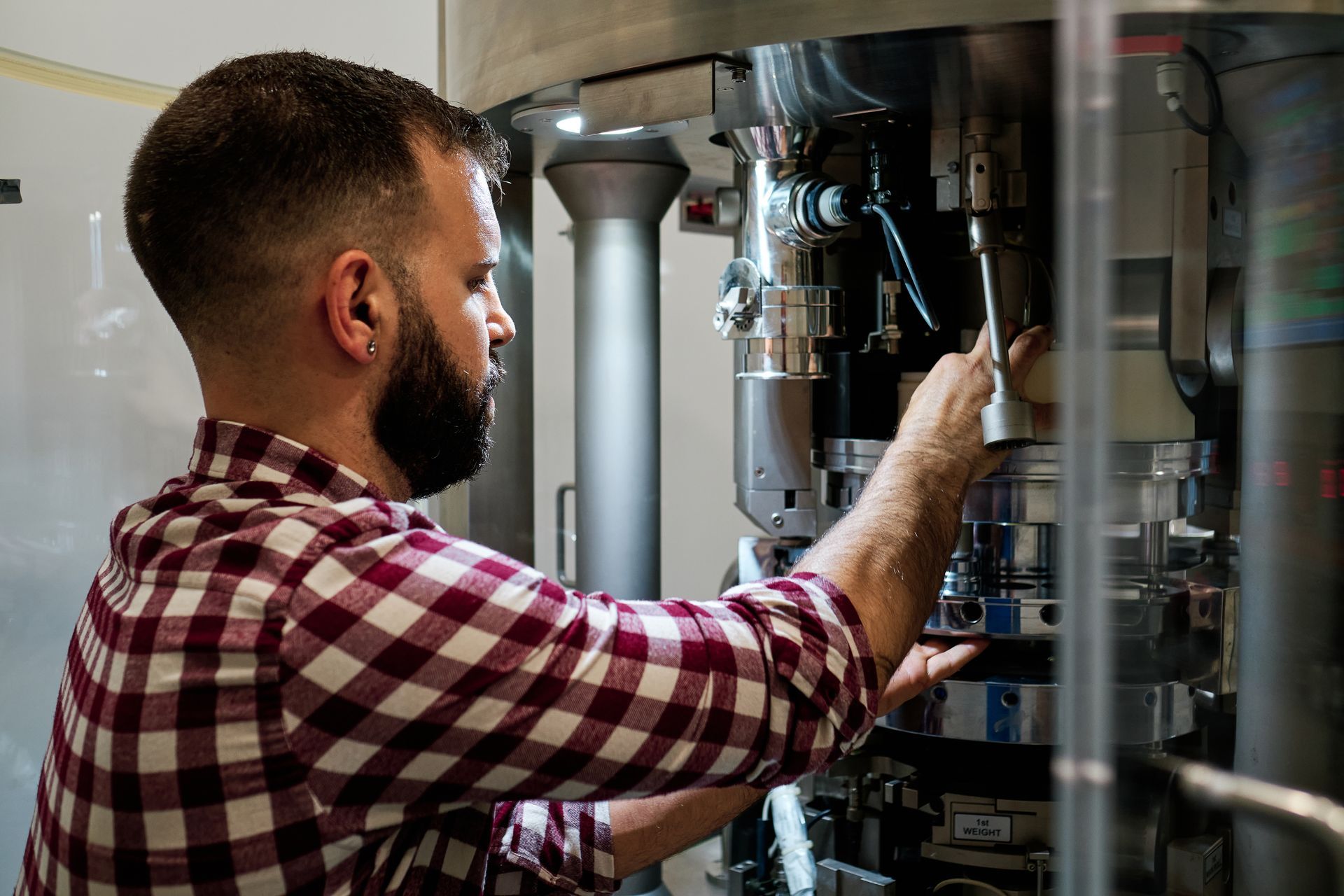 Man inspecting brewery machinery through a glass panel, reaching toward stainless steel tanks