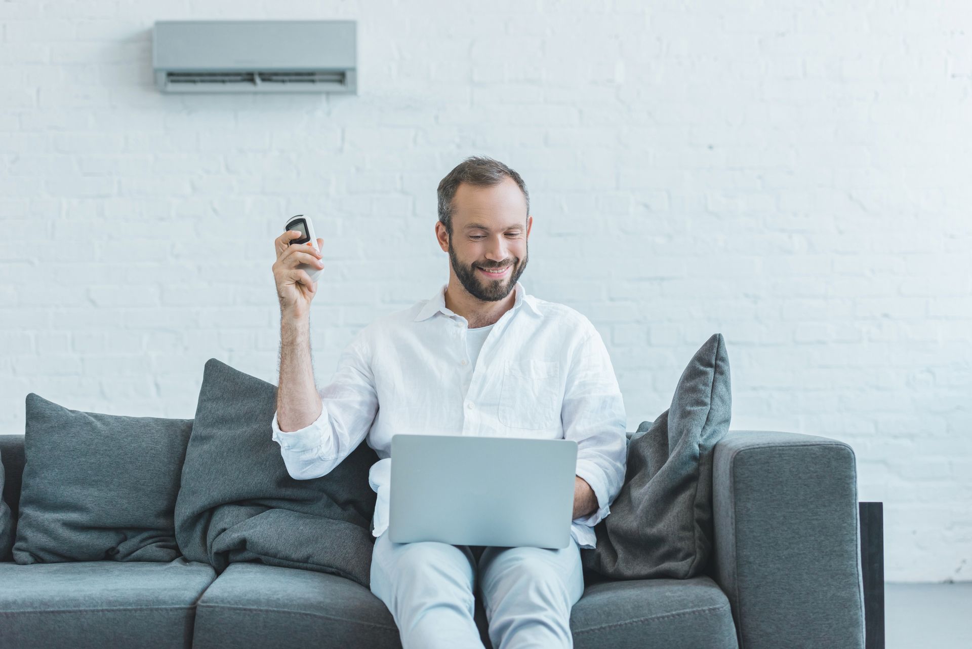 Man sitting on a gray couch using a laptop, holding a remote, in a bright living room with a wall-mounted air conditioner.