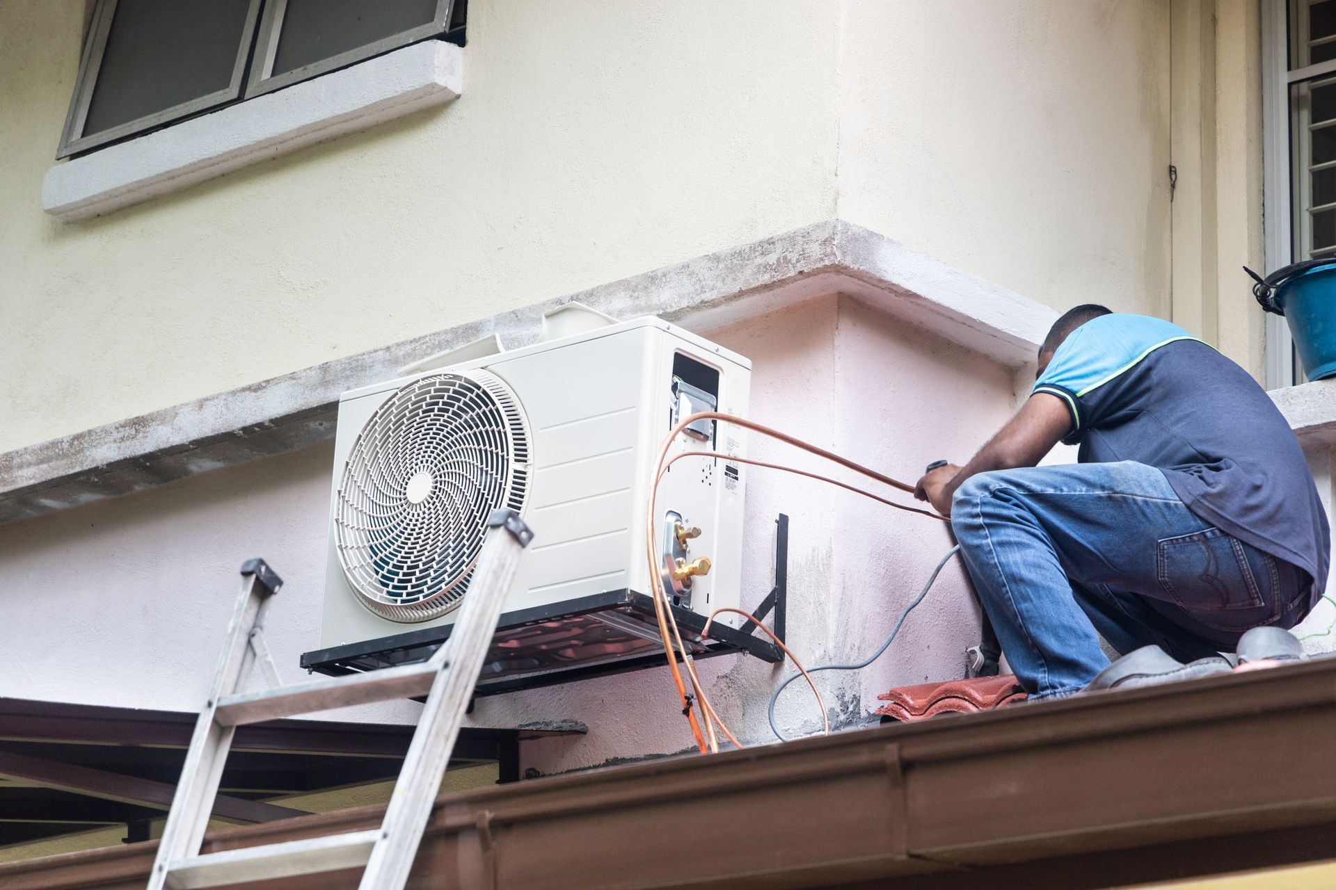 Technician repairing an outdoor air conditioner unit on a roof ledge with tools and cables.
