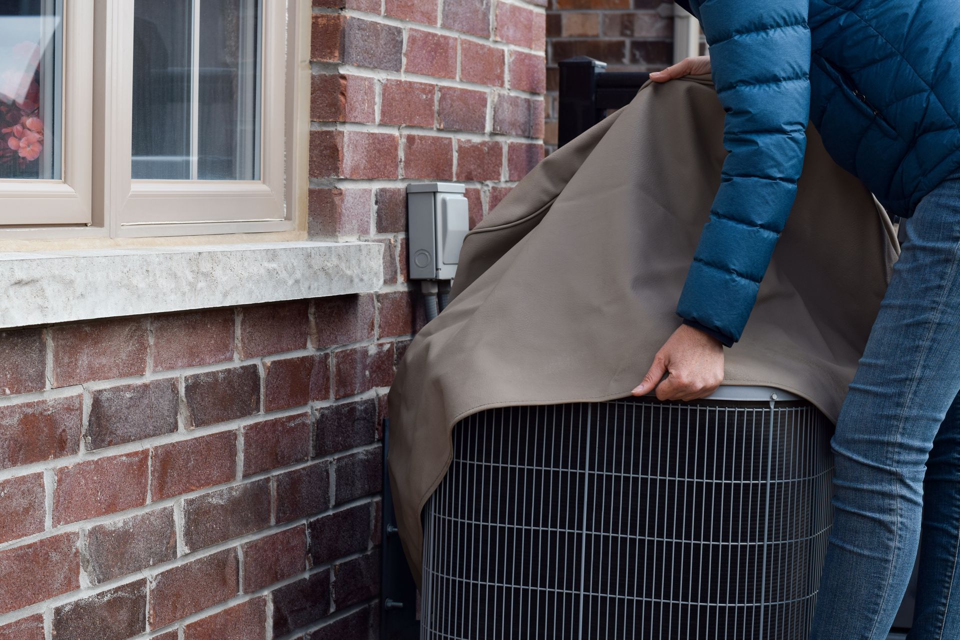 Person covering an outdoor air conditioner with a brown tarp beside a brick house