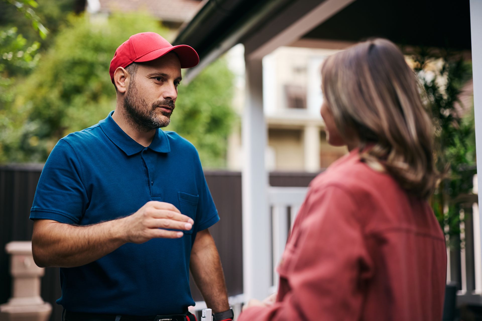 Man in red cap talking to woman at a doorstep, with a house and greenery in the background