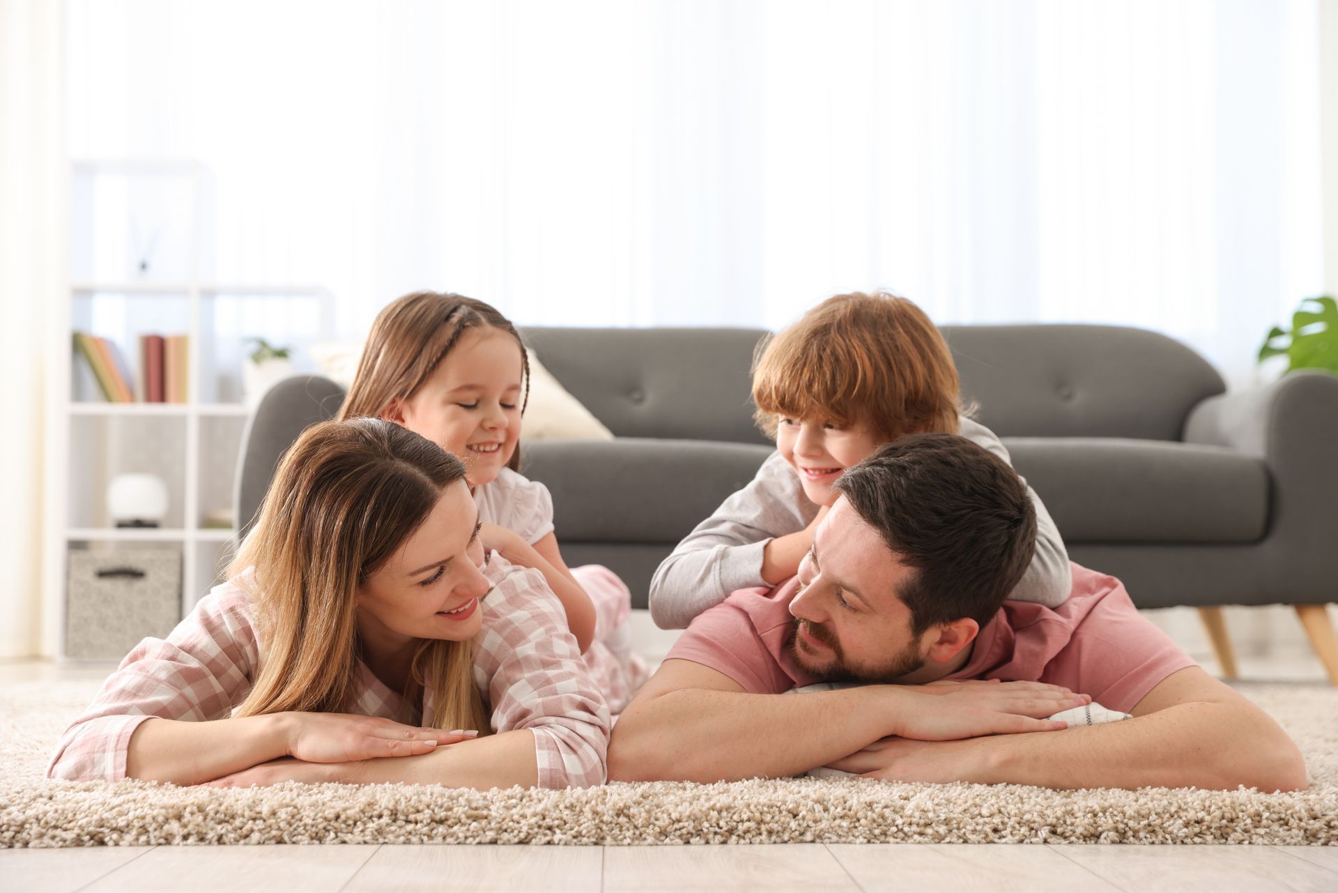 Family lying on a carpet in a bright living room, smiling and playing together.