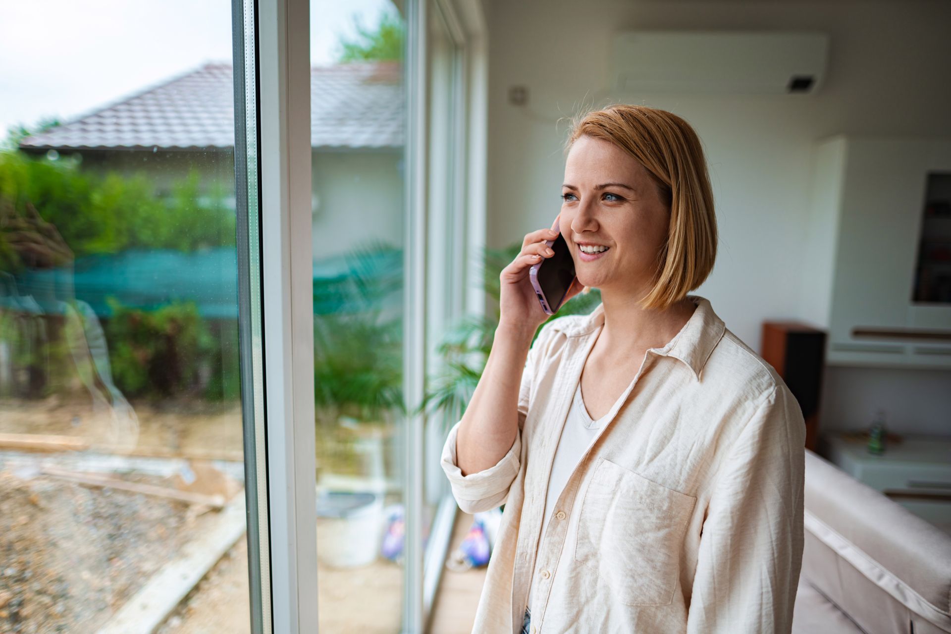 Woman smiling and talking on a phone by a large window in a bright home interior