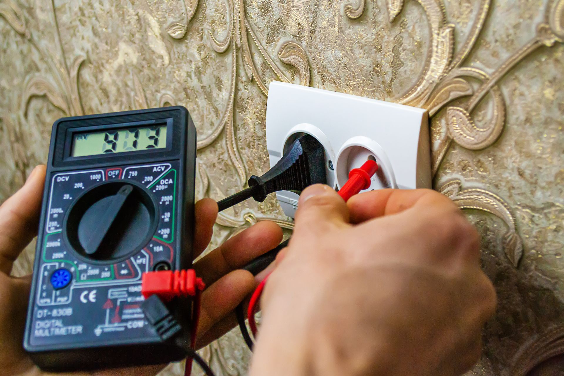 Hand holding multimeter probes against electrical outlet with wallpaper background