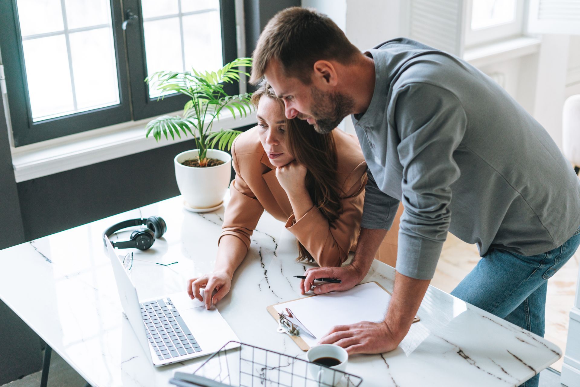 Two coworkers lean over a laptop at a bright desk, discussing work with notes and coffee nearby.