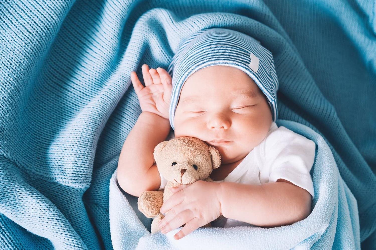 Sleeping baby wrapped in a blue blanket, holding a small teddy bear.