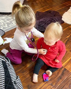 Two children on a hardwood floor, one using a toy stethoscope to examine the other.