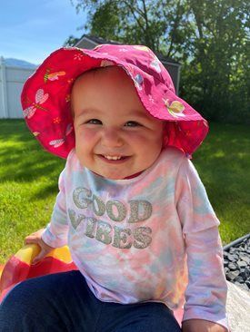 Smiling child in a pink sunhat and “GOOD VIBES” shirt sitting on grass outdoors