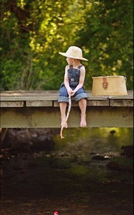 Child in sunhat sits on a wooden dock beside a basket above a dark pond