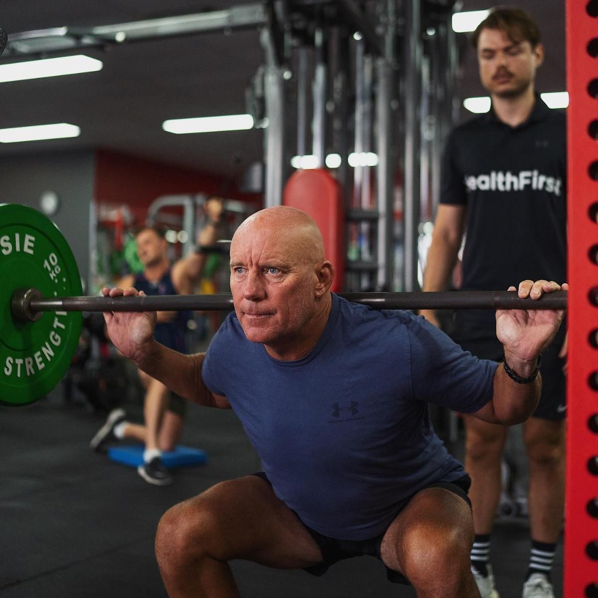 A Man is Squatting With a Barbell in a Gym — Health First Townsville In Townsville, QLD