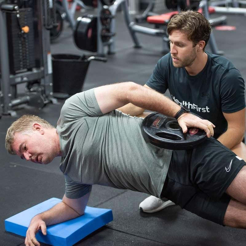 Two Men Are Doing Exercises in a Gym With One Wearing a Shirt That Says First — Health First Townsville In Townsville, QLD