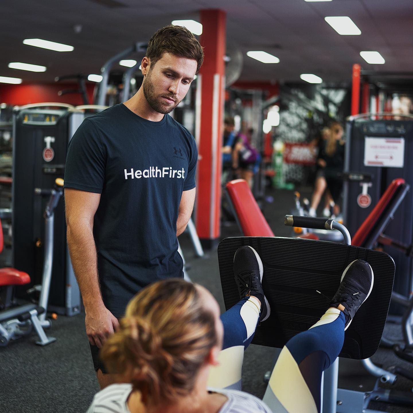 A Man Wearing a Black Shirt That Says Healthfirst Talks to a Woman in a Gym — Health First Townsville In Thuringowa, QLD