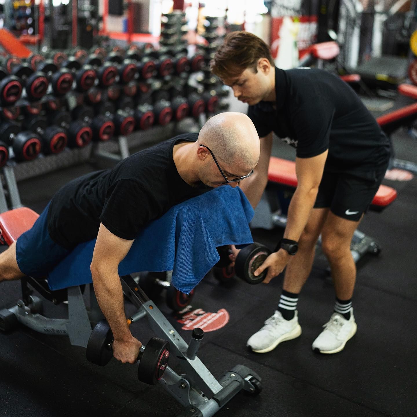A Man is Helping Another Man Lift a Dumbbell in a Gym — Health First Townsville In Townsville, QLD