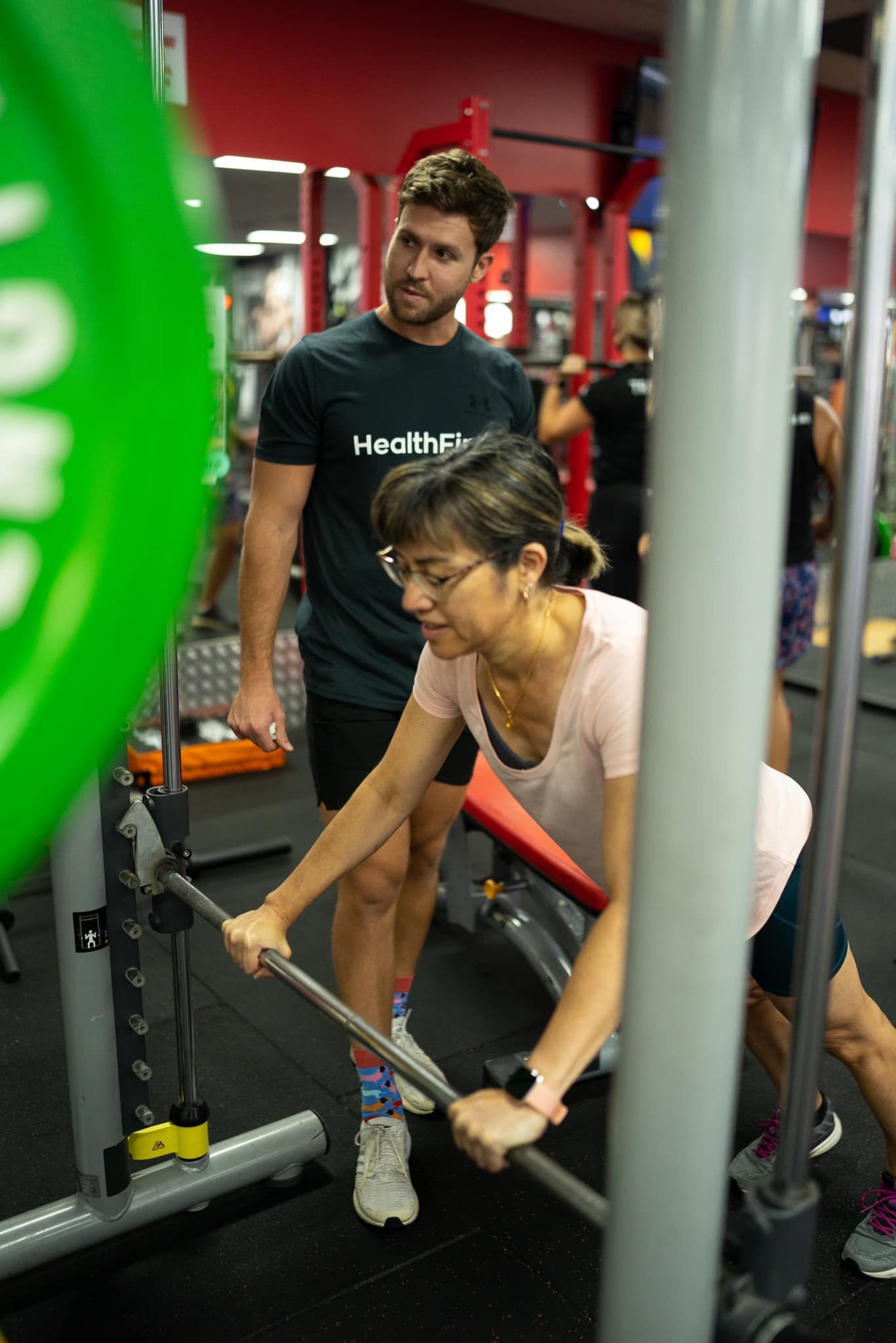 A Woman is Lifting a Barbell in a Gym While a Man Watches — Health First Townsville In Townsville, QLD