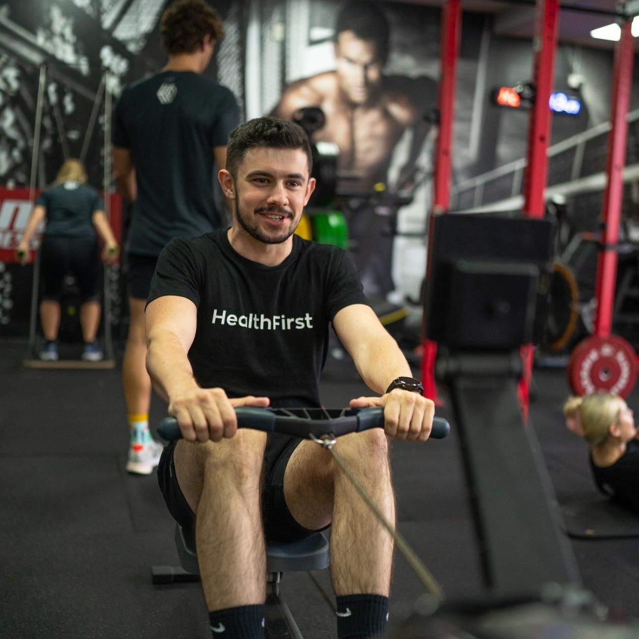 A Man Wearing a Black Shirt That Says Health First is Rowing in a Gym — Health First Townsville In Townsville, QLD
