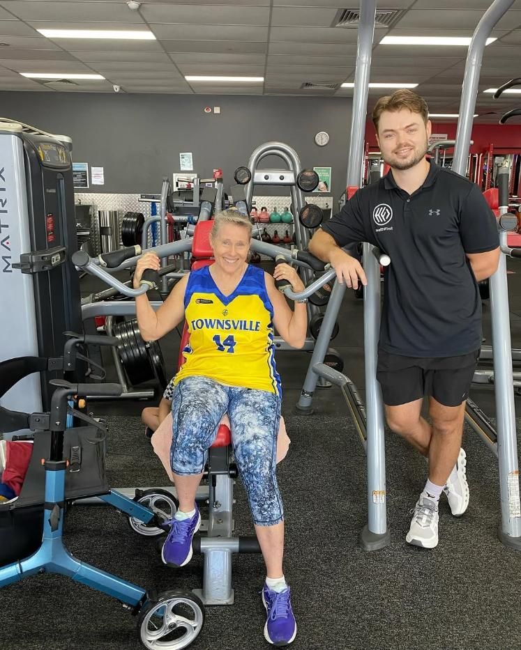 A Man is Standing Next to a Woman Sitting on a Machine in a Gym — Health First Townsville In Townsville, QLD