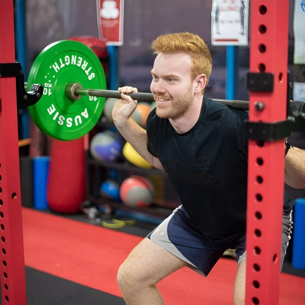 A Man Squatting With a Barbell That Says Aussie on It — Health First Townsville In Townsville, QLD