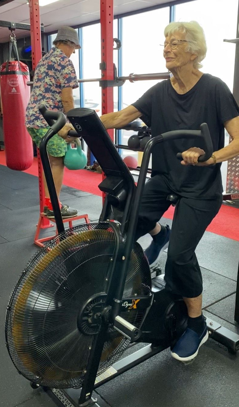 Woman exercising on an air bike at a gym, another person in the background.