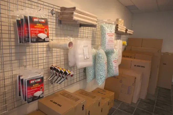 Store aisle with stacked cardboard boxes, hanging packaged items, and white protective suits on a wall rack.