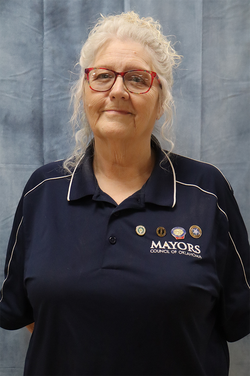 A woman wearing glasses and a blue mayors shirt is standing in front of a blue curtain.