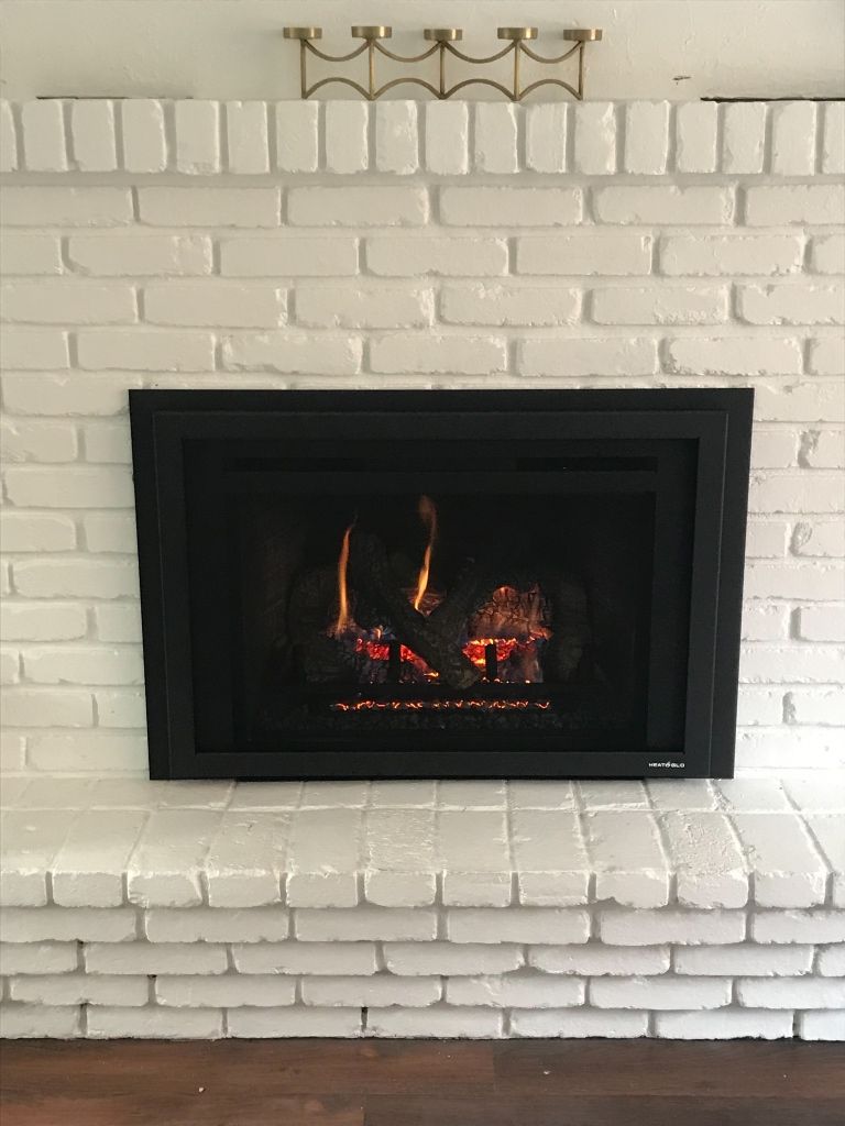 Fireplace with lit flames, framed by a black border, set in a white painted brick wall.