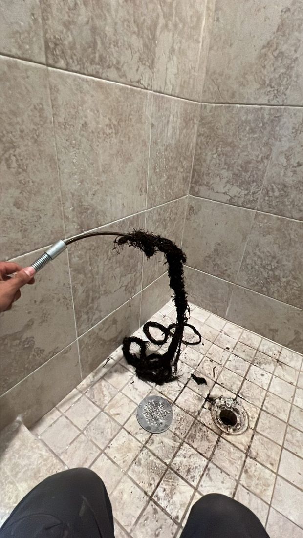 A drain snake pulls out a large clump of dark hair from a shower drain in a tile-walled shower.