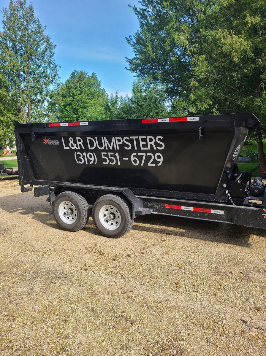 Black L&R Dumpsters trailer on gravel.  Phone number (319) 551-6729. Green trees and blue sky background.