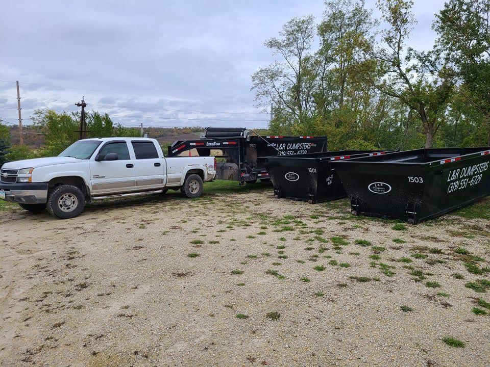 White pickup truck towing three black dumpsters on a gravel lot near trees.