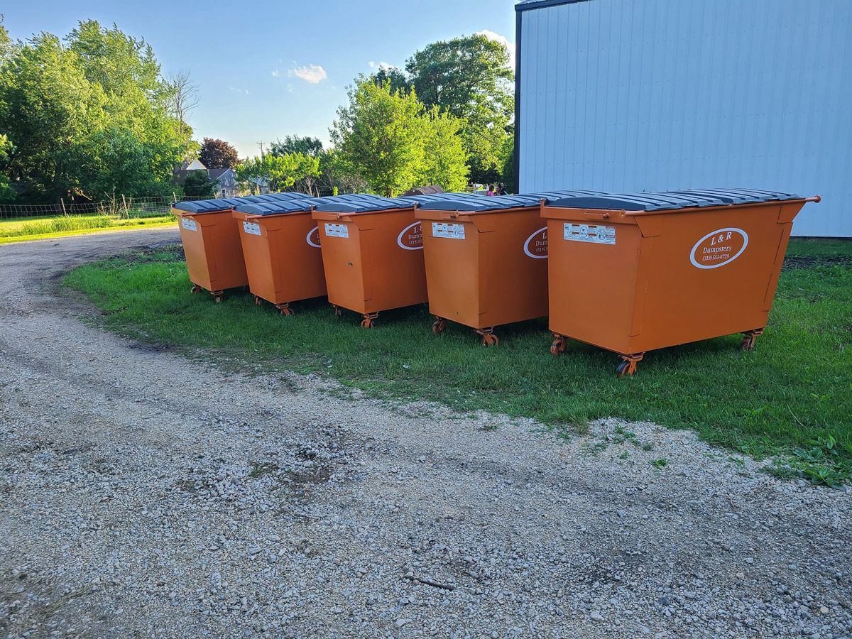 Six orange dumpsters lined up on grass near a gravel driveway and a white building.
