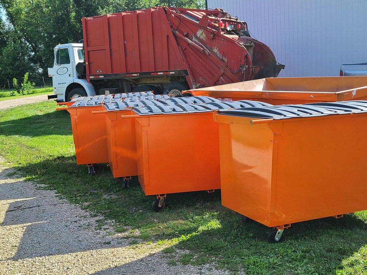Orange dumpsters on wheels with a red garbage truck in a grassy area.