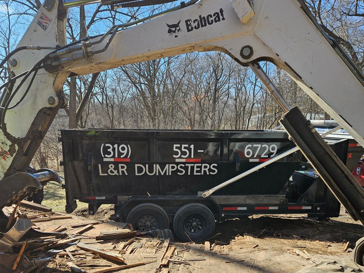 Bobcat excavator loading debris into L&R Dumpsters dumpster trailer; outdoors.