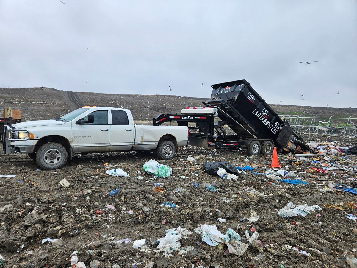 White pickup truck with a black dump trailer dumping trash at a landfill.