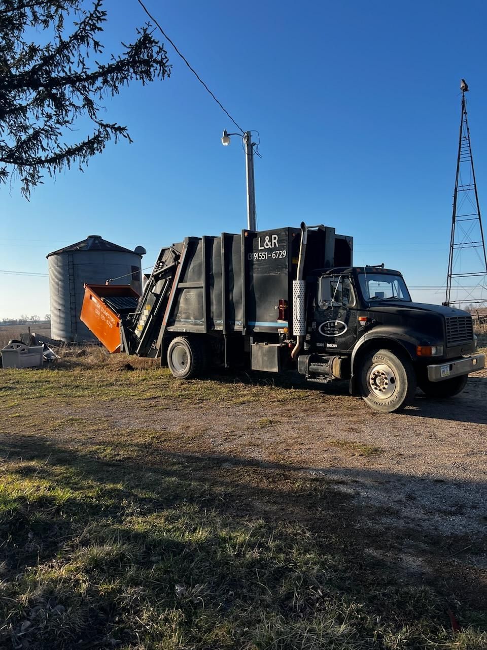 Black garbage truck on a grassy field; silo and utility pole in the background.