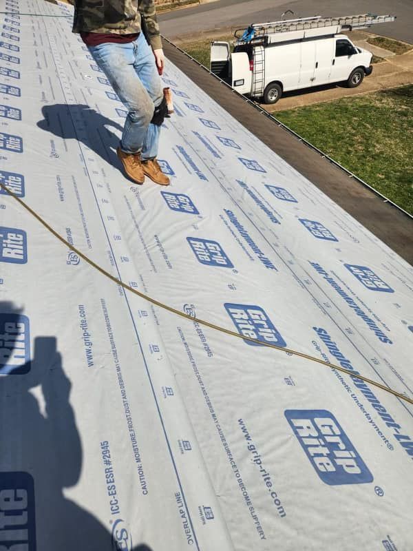 Person standing on a roof covered in Grip Rite underlayment, holding a tool. A white van is parked nearby.