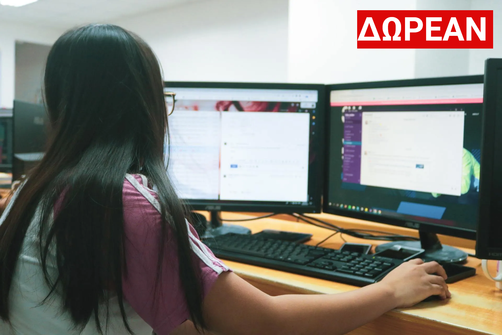 Γυναίκα με μακριά μαύρα μαλλιά χρησιμοποιεί υπολογιστή με δύο οθόνες, πληκτρολογεί σε πληκτρολόγιο και κάνει κλικ με το ποντίκι.
