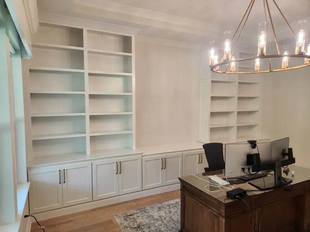 A home office with a desk , computer , shelves and a chandelier.