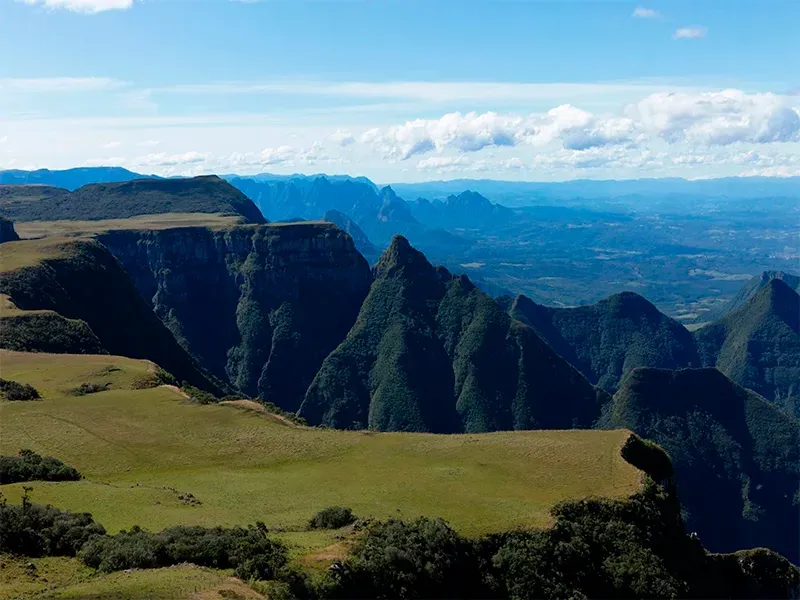 Vista panorâmica de um desfiladeiro com campos verdejantes, picos irregulares e um céu azul com nuvens.