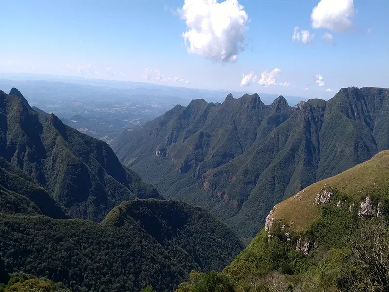 Cadeia montanhosa com encostas verdes e arborizadas sob um céu azul com algumas nuvens.