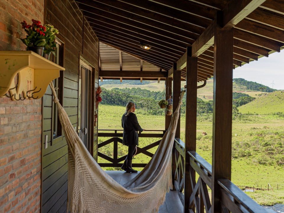 Mulher em pé numa varanda de madeira, olhando para uma paisagem verde, rede, decoração rústica.